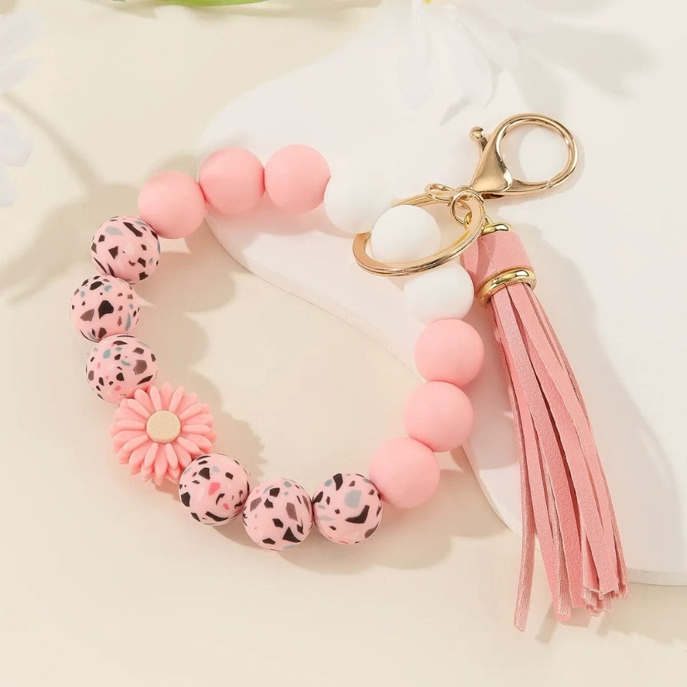 Assorted floral and daisy printed silicone bead bracelet keychains in pink, yellow, green, purple, brown, white, and black colorways with tassel accents displayed on a cream surface. - Pink