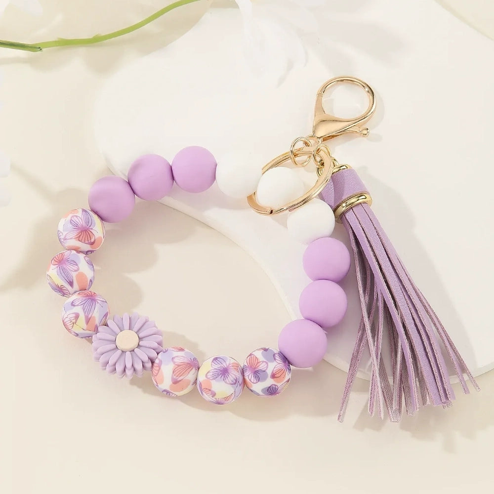 Assorted floral and daisy printed silicone bead bracelet keychains in pink, yellow, green, purple, brown, white, and black colorways with tassel accents displayed on a cream surface. - Purple