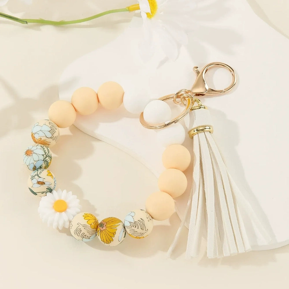 Assorted floral and daisy printed silicone bead bracelet keychains in pink, yellow, green, purple, brown, white, and black colorways with tassel accents displayed on a cream surface. - White