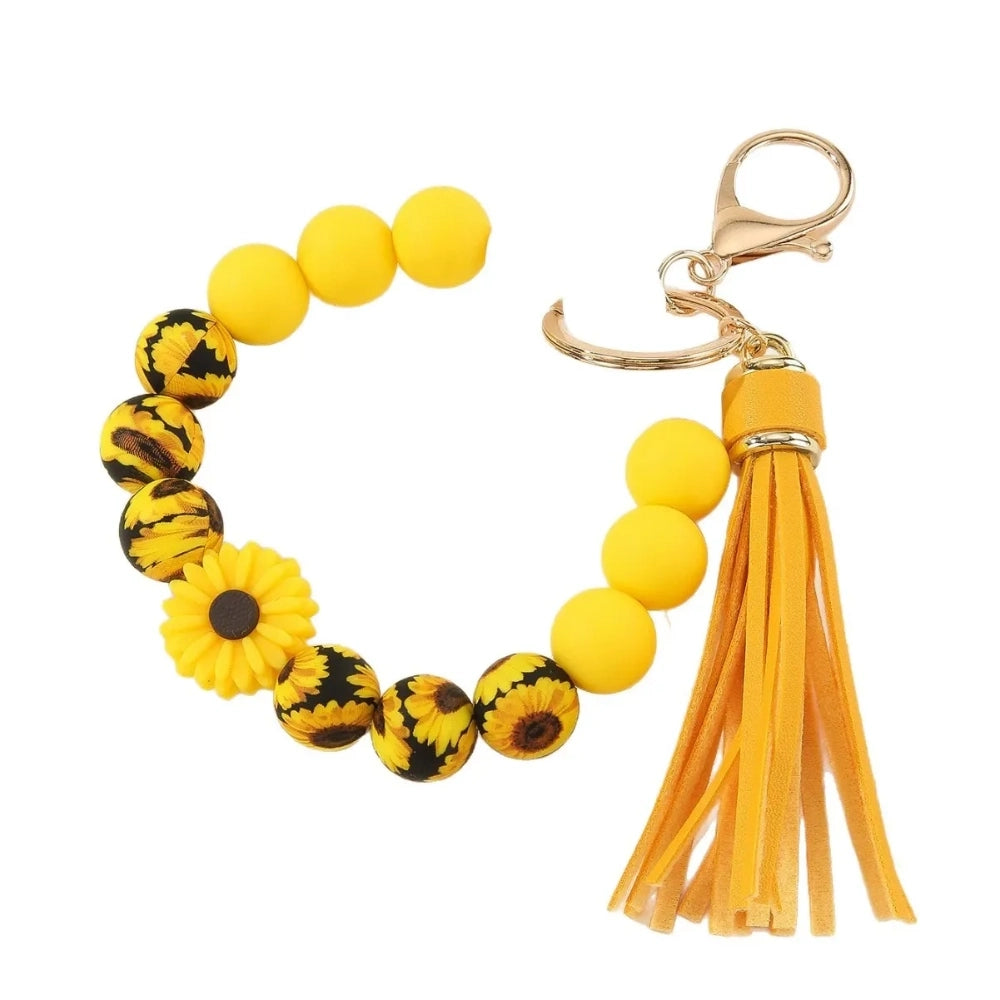 Assorted floral and daisy printed silicone bead bracelet keychains in pink, yellow, green, purple, brown, white, and black colorways with tassel accents displayed on a cream surface. - Yellow