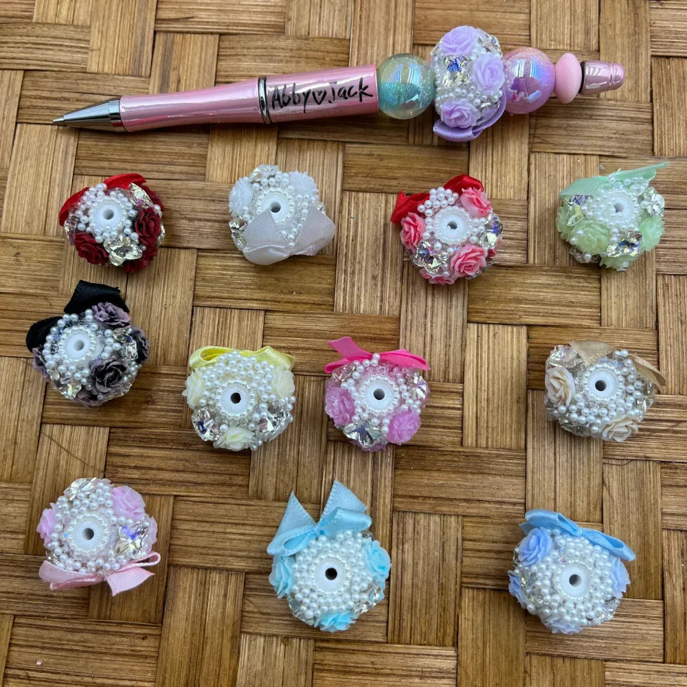 Assorted floral fancy beads decorated with pearl clusters, rhinestones, satin ribbon bows, and miniature roses in multiple colors including pink, blue, white, black, and green, displayed next to a pink beadable pen on a wicker surface.