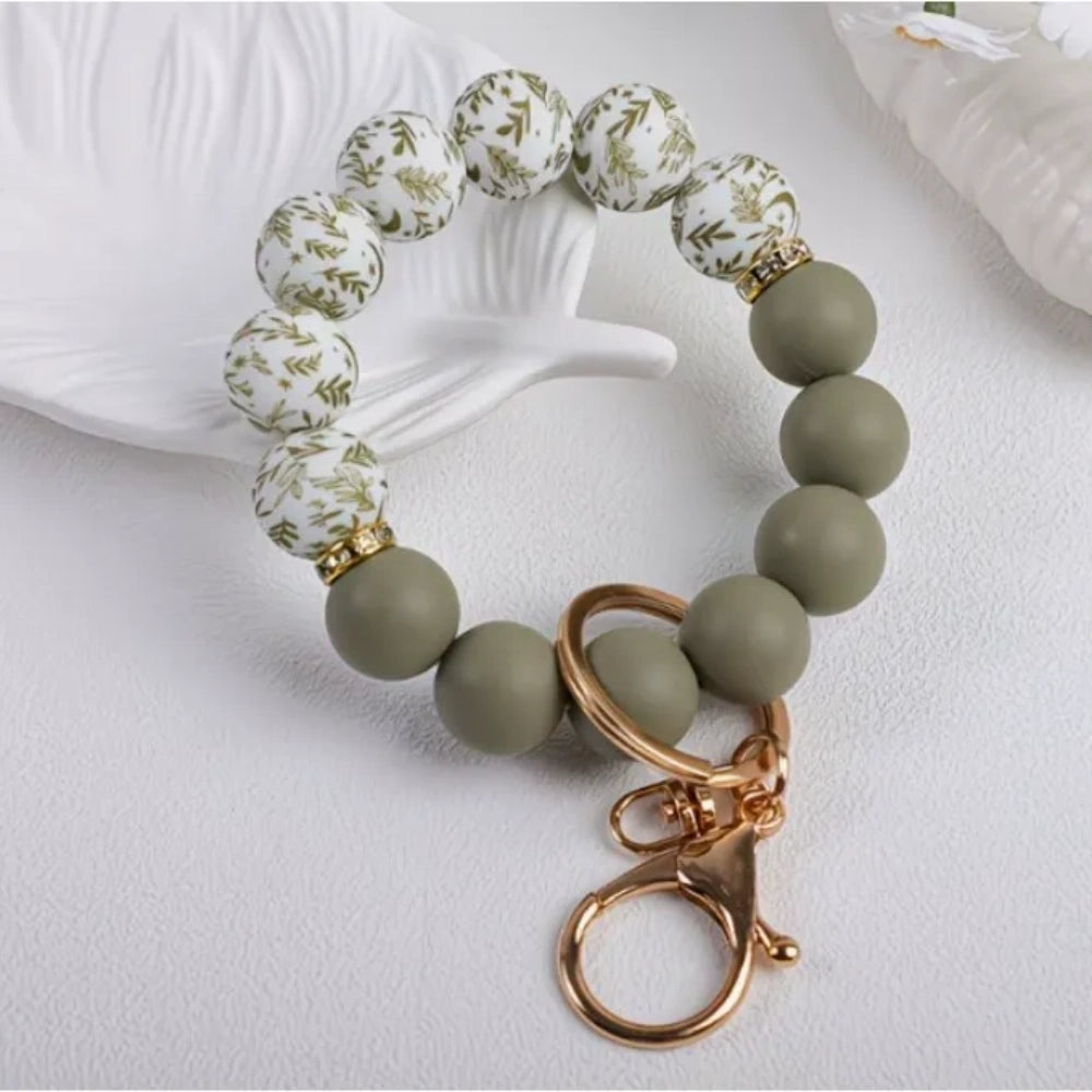 Assorted floral print and solid matte silicone bead bracelets with gold lobster clasp keychains in black, pink, sage green, and gray colorways displayed on a white round tray with white flowers in background. - 1