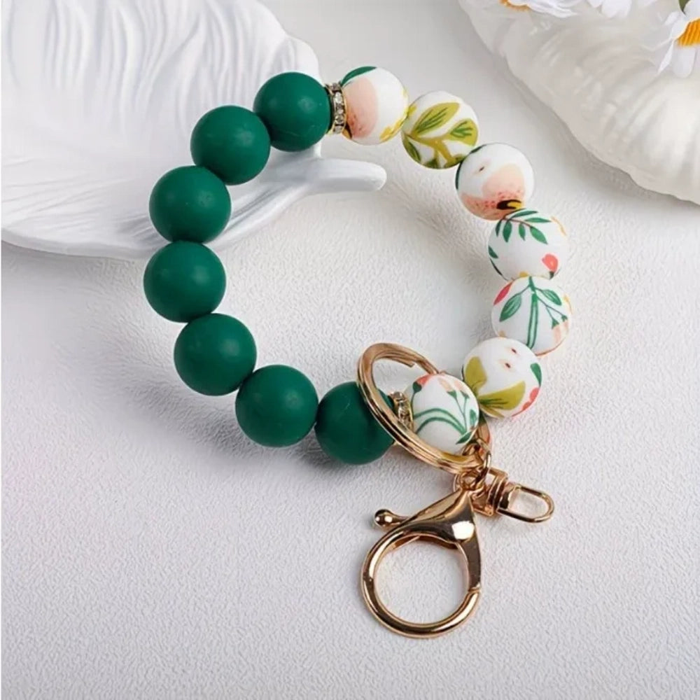 Assorted floral print and solid matte silicone bead bracelets with gold lobster clasp keychains in black, pink, sage green, and gray colorways displayed on a white round tray with white flowers in background. - 2