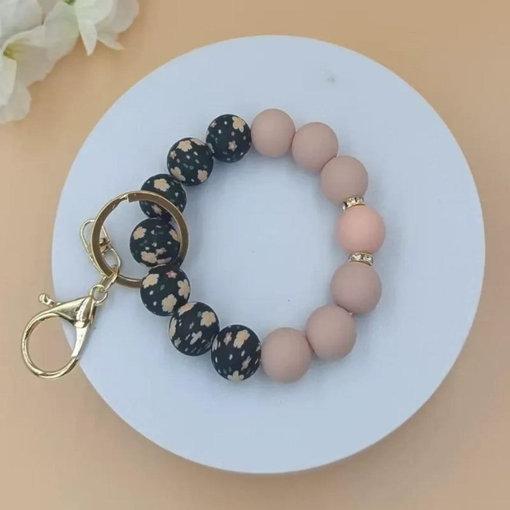 Assorted floral print and solid matte silicone bead bracelets with gold lobster clasp keychains in black, pink, sage green, and gray colorways displayed on a white round tray with white flowers in background. - 3