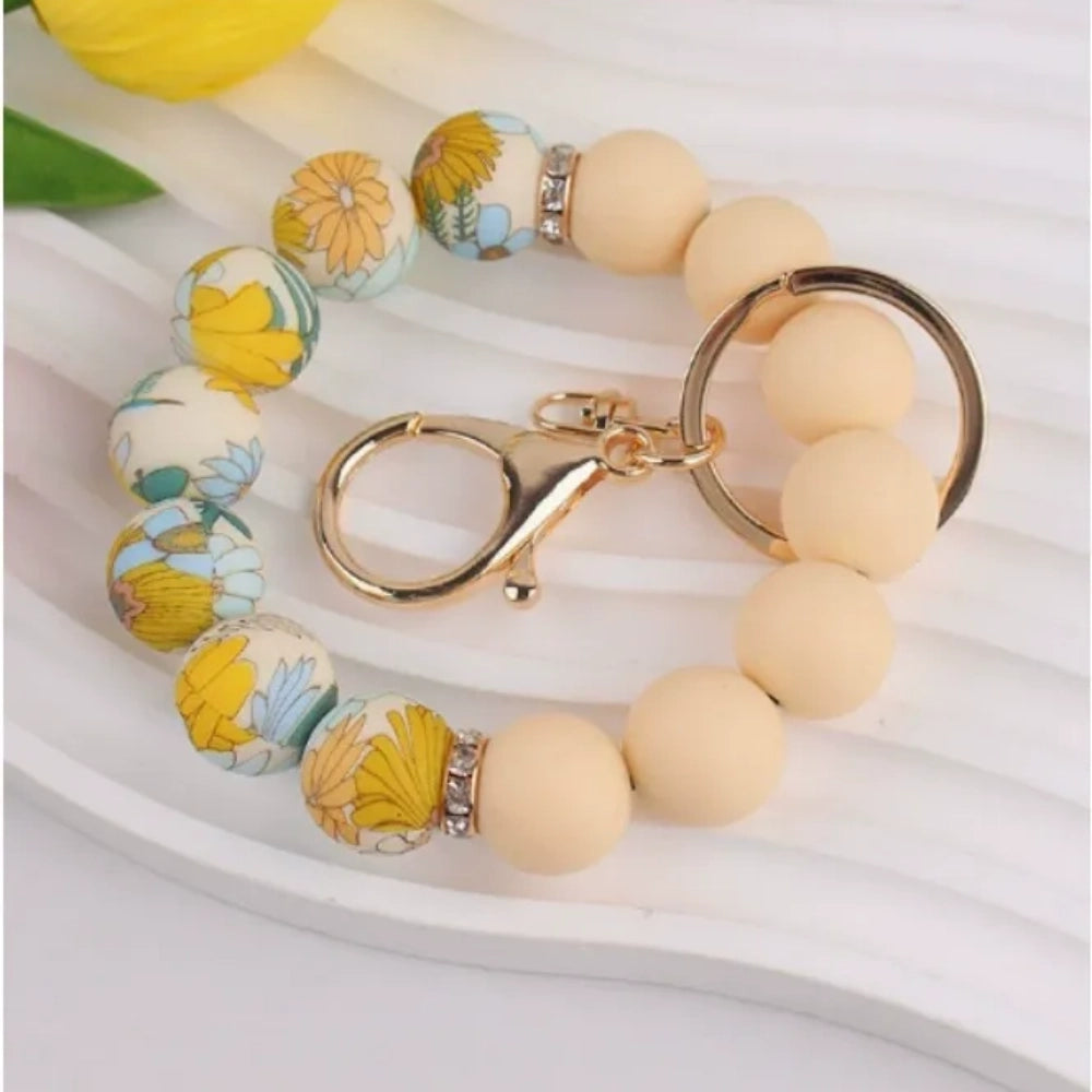 Assorted floral print and solid matte silicone bead bracelets with gold lobster clasp keychains in black, pink, sage green, and gray colorways displayed on a white round tray with white flowers in background. - 4