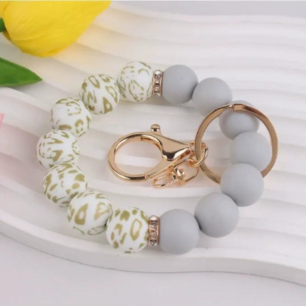 Assorted floral print and solid matte silicone bead bracelets with gold lobster clasp keychains in black, pink, sage green, and gray colorways displayed on a white round tray with white flowers in background. - 5