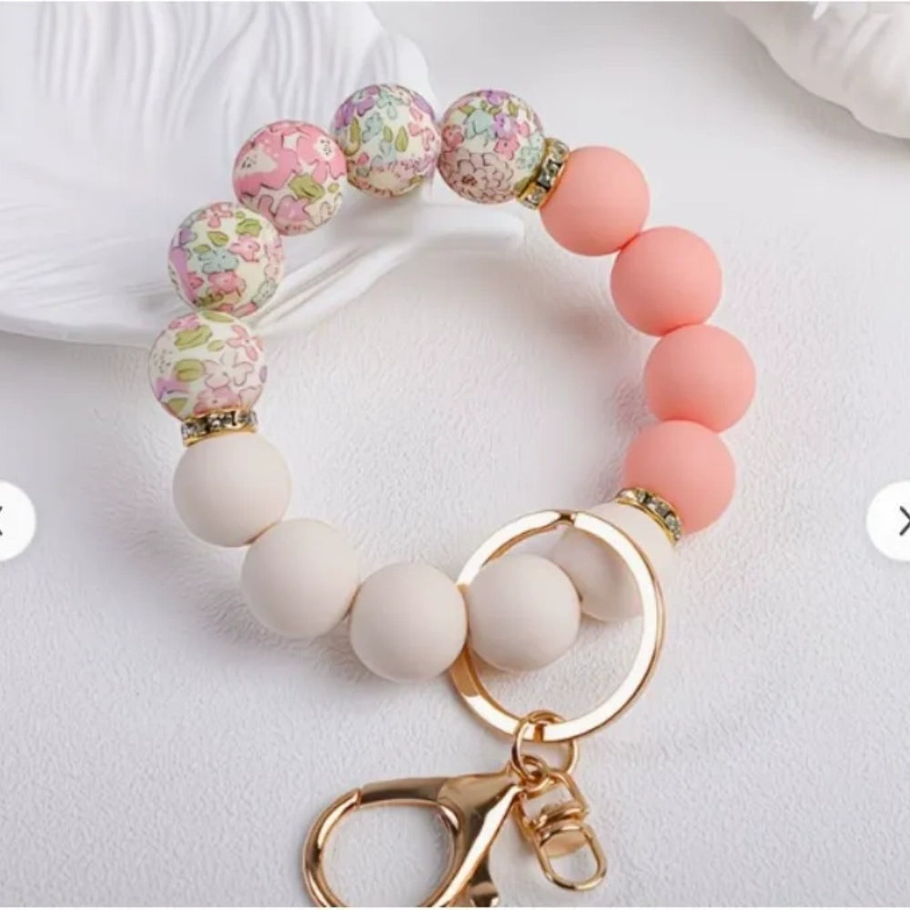 Assorted floral print and solid matte silicone bead bracelets with gold lobster clasp keychains in black, pink, sage green, and gray colorways displayed on a white round tray with white flowers in background. - 6