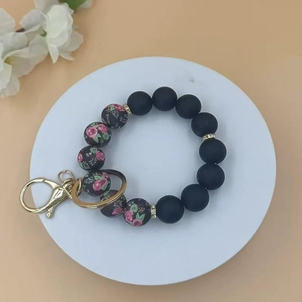 Assorted floral print and solid matte silicone bead bracelets with gold lobster clasp keychains in black, pink, sage green, and gray colorways displayed on a white round tray with white flowers in background. - 7