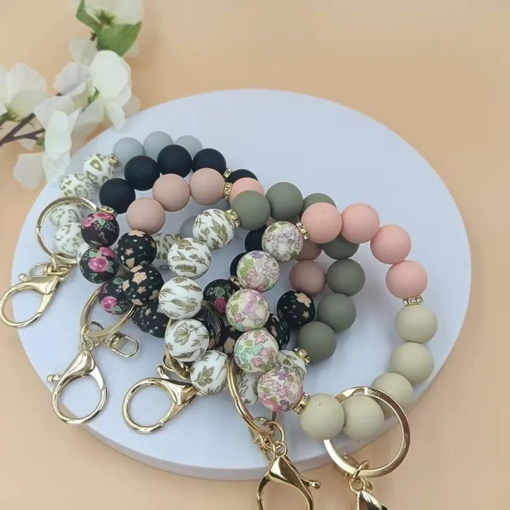 Assorted floral print and solid matte silicone bead bracelets with gold lobster clasp keychains in black, pink, sage green, and gray colorways displayed on a white round tray with white flowers in background. - Random Mixed