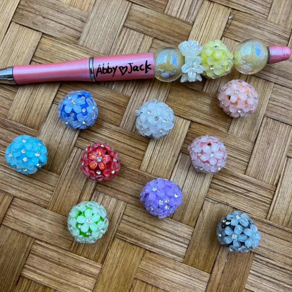 Assorted colorful floral rhinestone acrylic fancy beads in blue, red, purple, green, pink, white, and gray, displayed next to a pink beadable pen labeled Abby and Jack on a woven rattan surface.