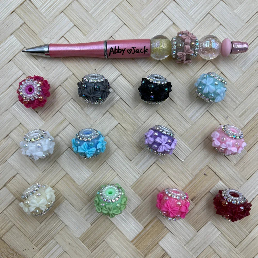 Assorted floral rhinestone fancy beads in multiple colors including pink, red, black, gray, white, blue, purple, green and burgundy, displayed next to a pink beadable pen on a woven bamboo surface.