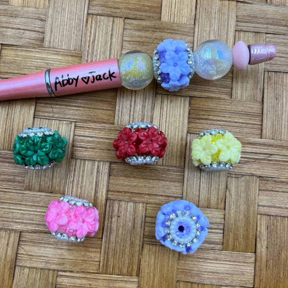 Colorful floral rhinestone fancy beads in green, red, yellow, pink, and blue with silver rhinestone trim, displayed with a pink beadable pen on a wicker surface.