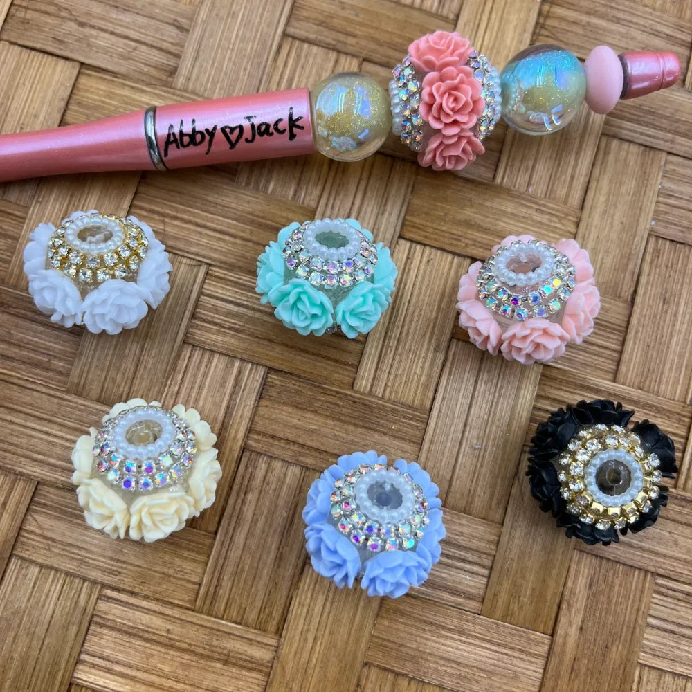 Assorted floral rhinestone pearl fancy beads in white, mint, pink, cream, blue, and black with crystal center accents, displayed on a woven surface alongside a decorated beadable pen.