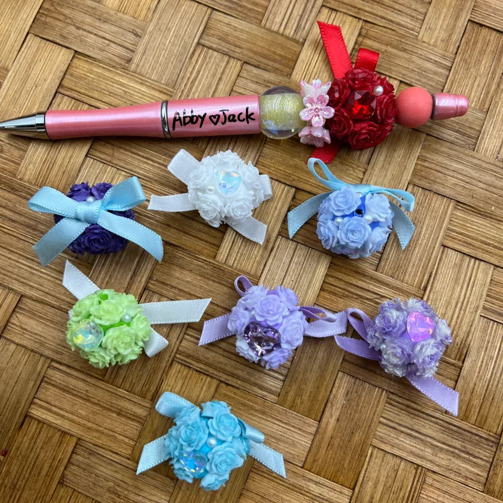 Assorted colorful floral rose ball fancy beads with satin ribbon bows and rhinestone gems in purple, white, blue, green, lavender, and red, displayed on a woven surface alongside a decorated beadable pen labeled Abby and Jack.