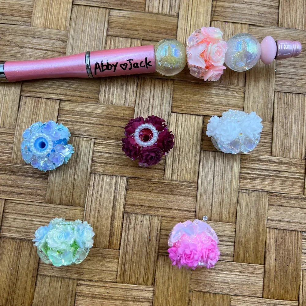 Assorted floral rose cluster resin fancy beads in pink, burgundy, white, blue, and green with iridescent shimmer, displayed alongside a custom beadable pen on a woven bamboo surface.