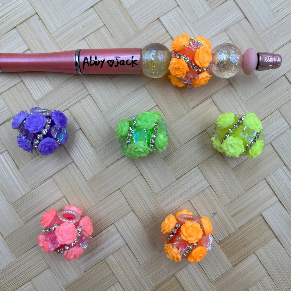 Colorful floral rose rhinestone fancy beads in purple, green, yellow-green, pink, and orange with butterfly accents, shown with a decorated beadable pen on a woven background.
