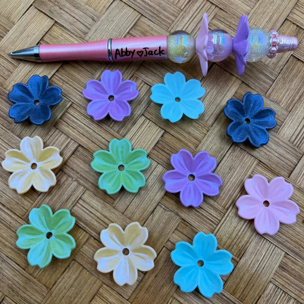 Assorted color flower-shaped acrylic spacer beads in blue, purple, teal, green, pink, and cream displayed next to a beadable pen on a woven surface.