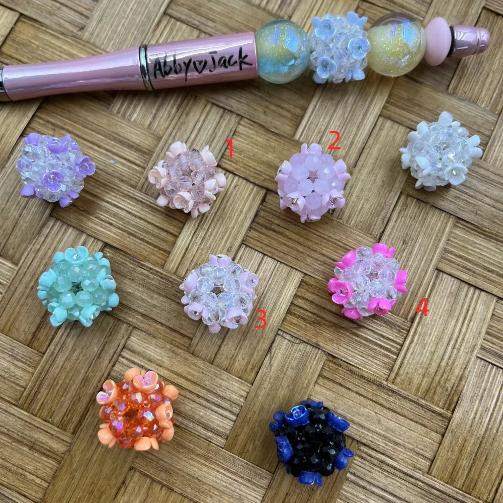 Assorted flower cluster crystal fancy beads in multiple colors including lavender, pink, mint, white, hot pink, orange, and navy blue arranged on a woven surface next to a decorated beadable pen labeled Abby and Jack.