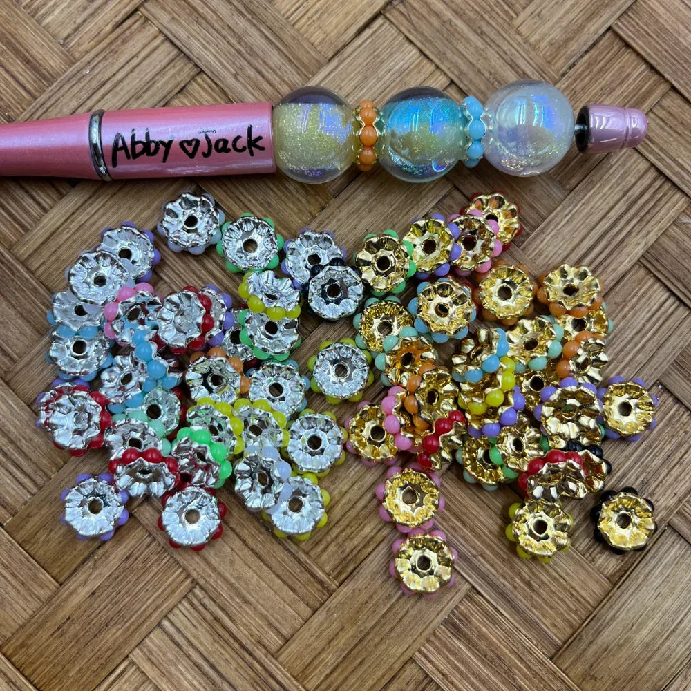 Assorted silver and gold flower daisy metal spacer beads with multicolor pastel petals displayed on woven bamboo surface next to a decorated beadable pen labeled Abby and Jack.