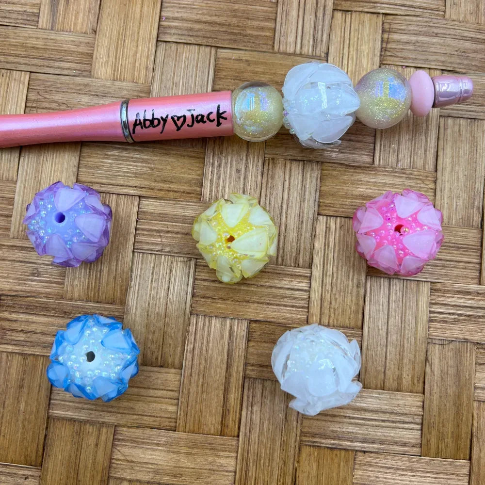 Assorted pastel flower petal rhinestone chunky beads in purple, yellow, pink, blue, and white displayed on a woven rattan surface alongside a pink beadable pen decorated with iridescent and floral beads labeled Abby and Jack.