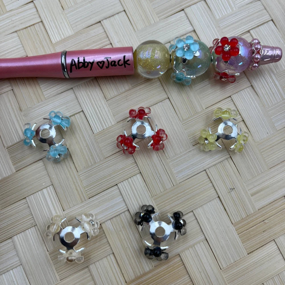 Assorted colorful flower petal bail beads in blue, red, yellow, clear, and black with silver metal frames, shown with a pink beadable pen labeled Abby Jack on a woven bamboo background.