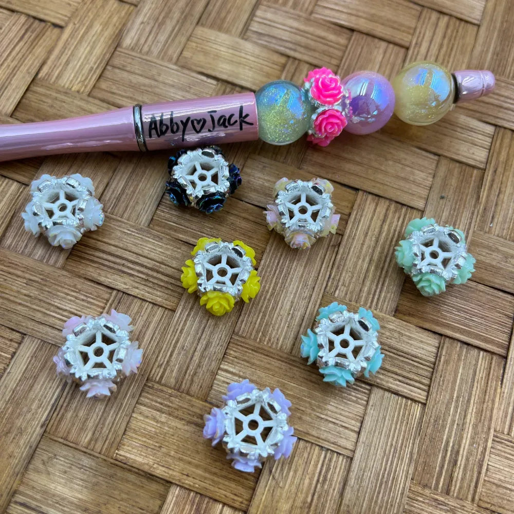 Assorted pastel flower rose acrylic spacer beads with silver filigree frame in white, yellow, mint, lavender, and dark blue, displayed next to a decorated beadable pen on a wicker surface.