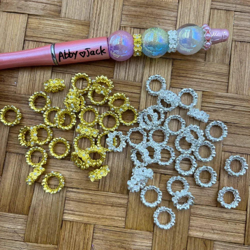Gold and silver flower wreath metal spacer rings displayed next to a decorated beadable pen on a wicker surface, craft supplies for DIY jewelry making.