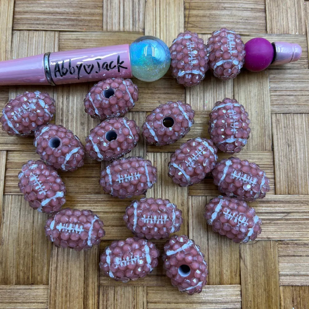 Brown football-shaped rhinestone acrylic beads with white lace stitching detail, shown next to a pink beadable pen on a woven surface