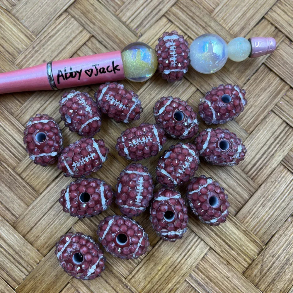 Brown and white football-shaped rhinestone beads with lace stitch detail, shown next to a beadable pen on a wicker background.