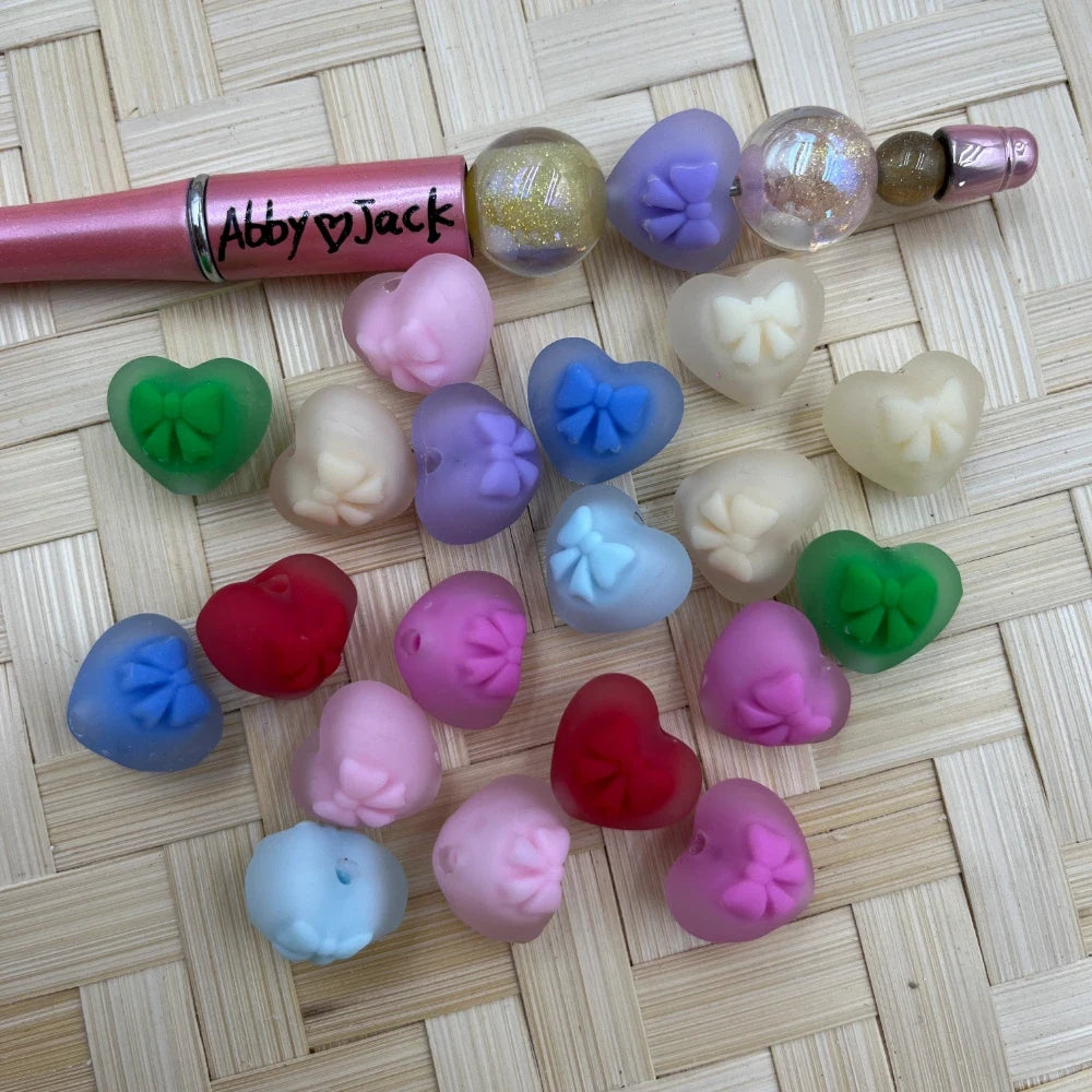Assorted frosted heart-shaped acrylic beads with raised bow detail in multiple colors including pink, purple, blue, green, red, and cream, displayed on a woven surface next to a pink beadable pen labeled Abby Jack.