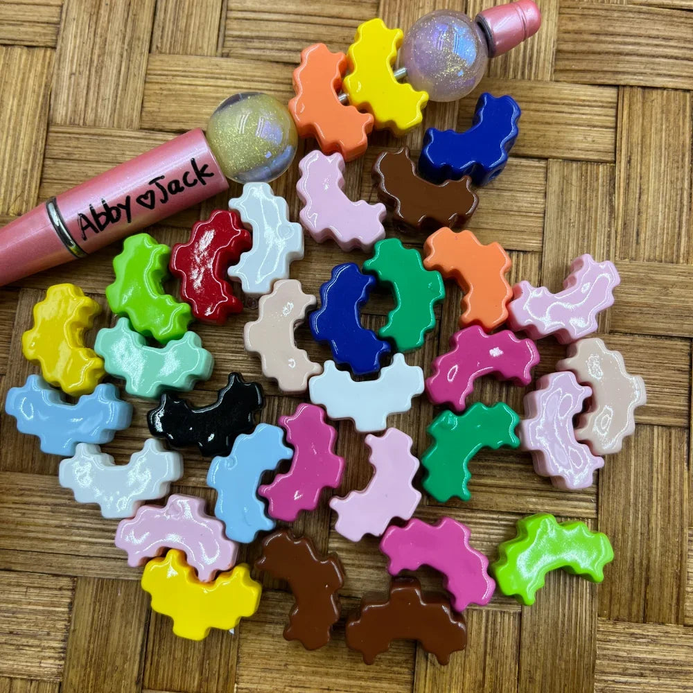 Assorted colorful glossy gear shaped acrylic beads in mixed colors including pink, blue, green, yellow, brown, black, and more, displayed on a wicker surface next to beadable pens.