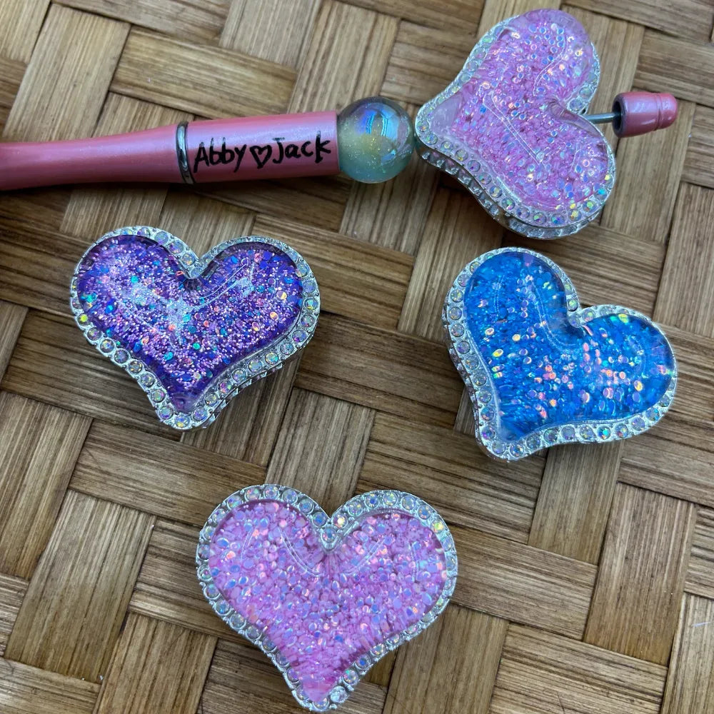 Four glitter-filled heart-shaped resin beads with rhinestone borders in pink, purple, and blue colors, displayed on a wicker surface alongside a beadable pen labeled Abby and Jack.