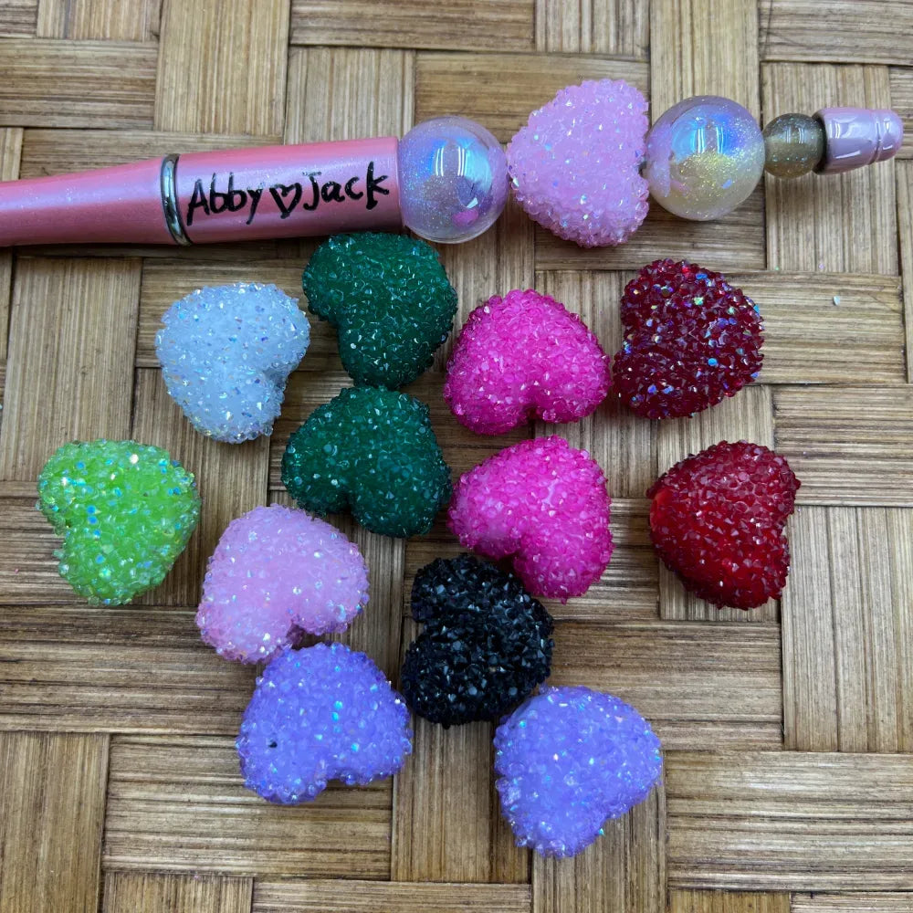 Assorted glitter heart-shaped sugar resin beads in pink, red, green, purple, black, white, and lime green colors displayed on a woven surface next to a pink beadable pen labeled Abby Jack.