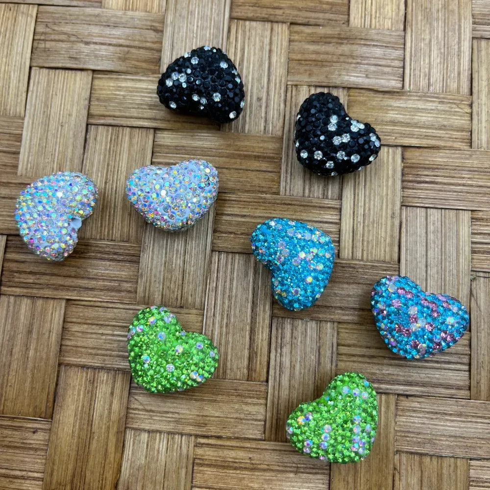 Glitter rhinestone heart-shaped beads in black, iridescent white, teal blue, and lime green displayed in pairs on a woven rattan surface for DIY craft projects.