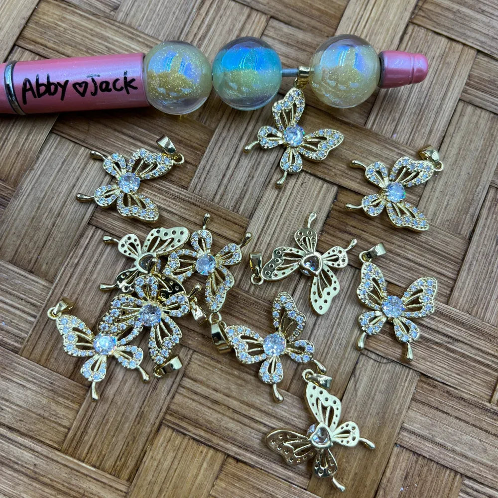 Gold butterfly charm bail beads with CZ crystal rhinestone wings and center stone, displayed on woven surface next to beadable pen components, for DIY jewelry making.