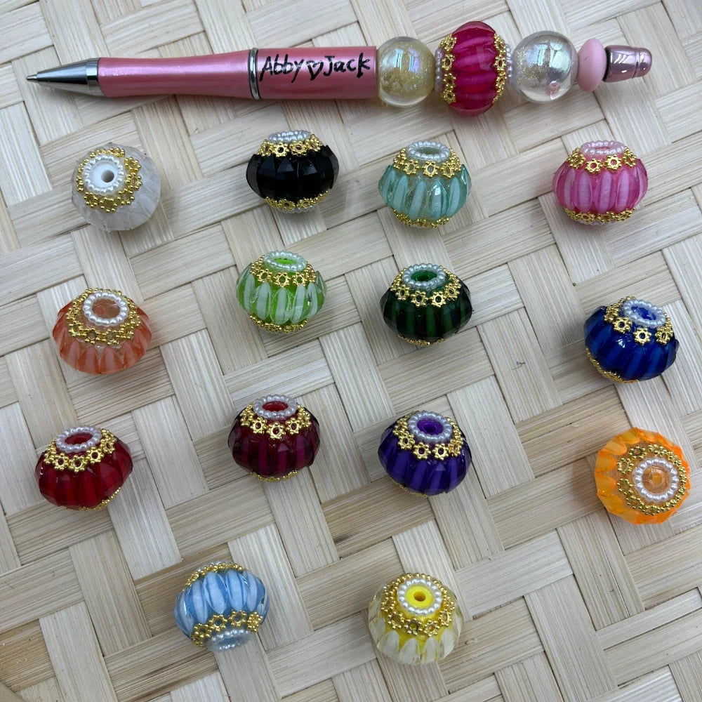 Assorted colorful ribbed fancy beads with gold daisy caps and pearl accents in 14 colors including red, blue, green, purple, orange, and pink, displayed on a woven surface next to a beadable pen.