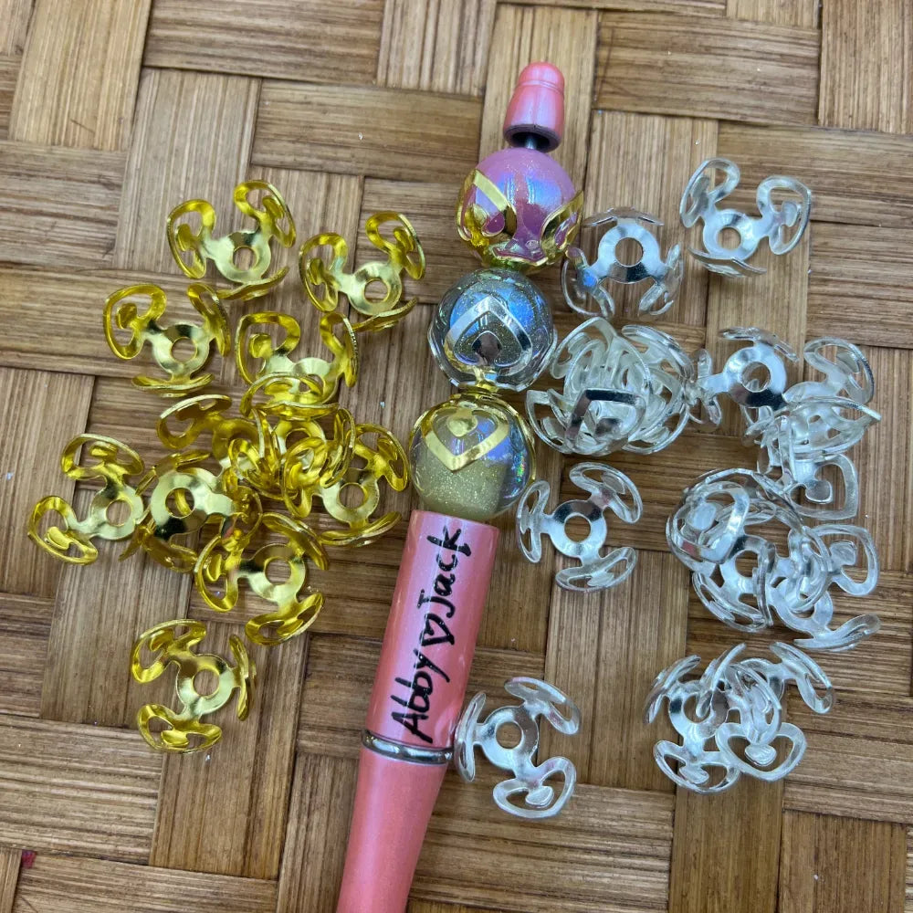 Gold and clear transparent flower-shaped plastic bead caps displayed next to a decorated pink beadable pen on a woven bamboo surface, used for DIY jewelry making, keychains, and badge reels.