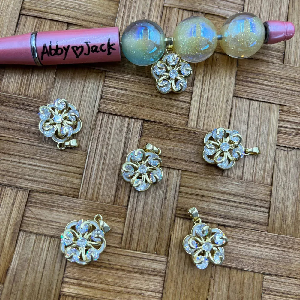 Gold metal flower-shaped bail beads with iridescent crystal rhinestone accents displayed on woven wood surface next to beadable pen and glitter globe beads