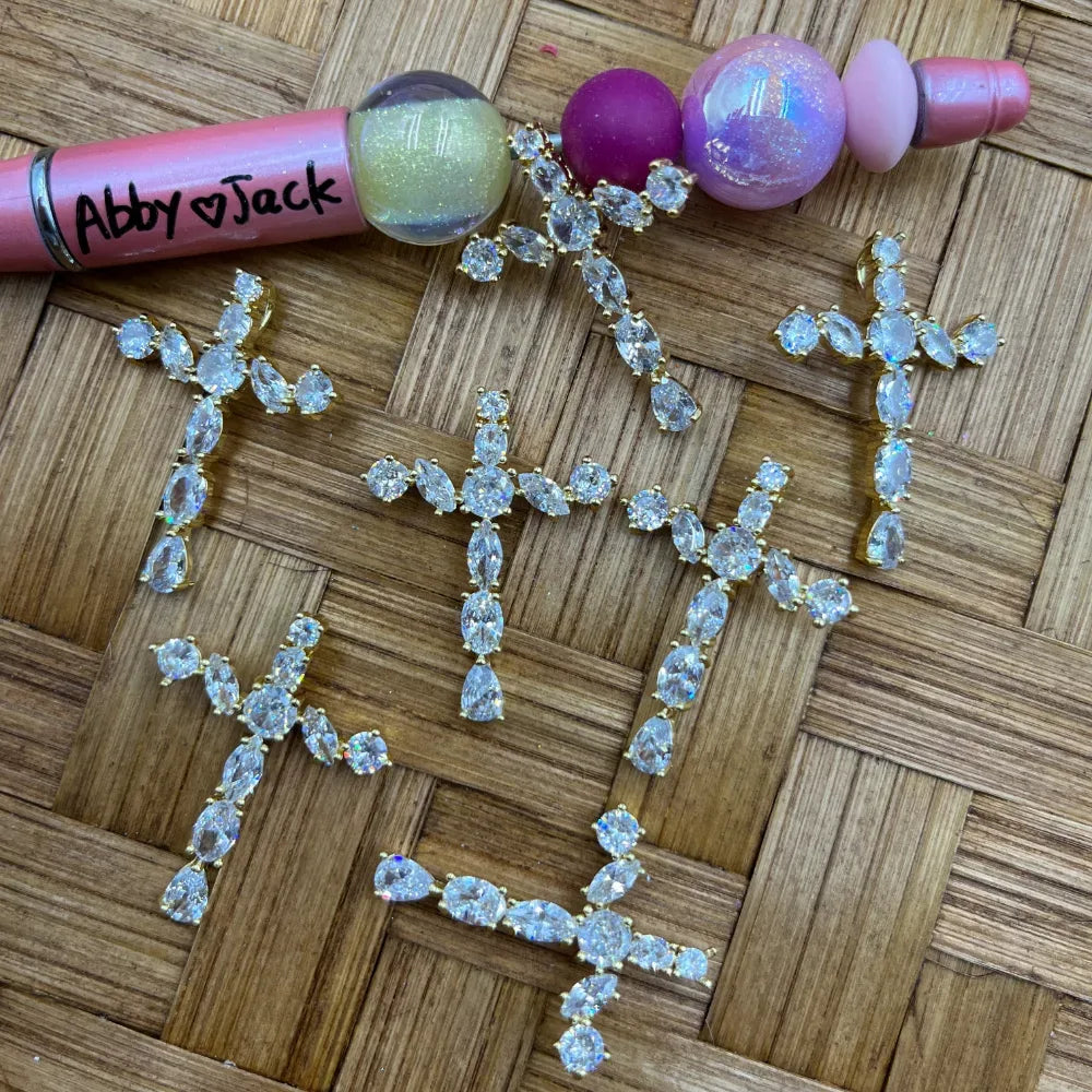 Gold-tone crystal cross rhinestone bail beads with marquise and round CZ stones displayed on woven bamboo surface next to beadable pens, showing multiple cross pendant charms for DIY jewelry making.