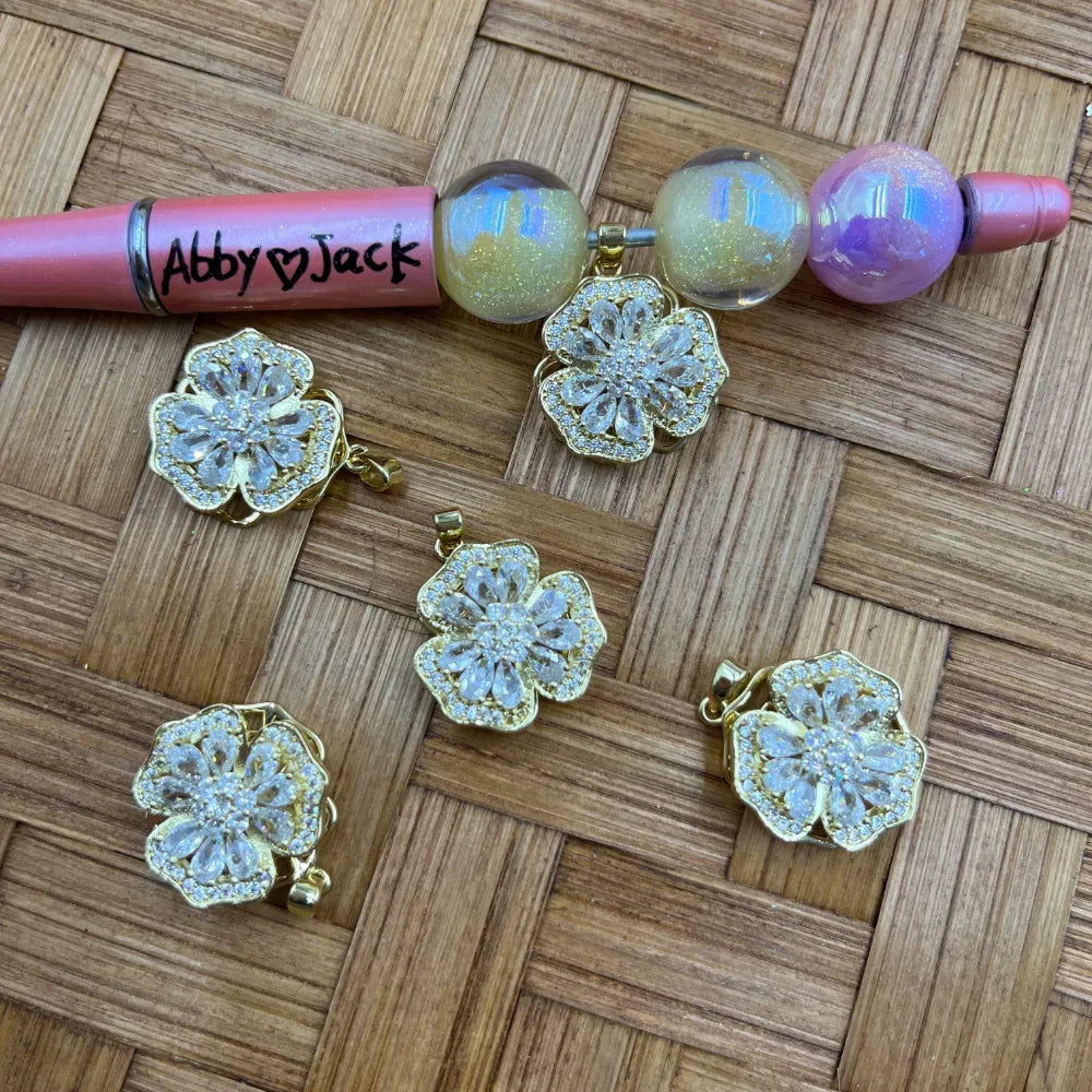 Gold cubic zirconia crystal flower bail beads with clear rhinestone petals and pave border, shown with a beadable pen on woven bamboo background