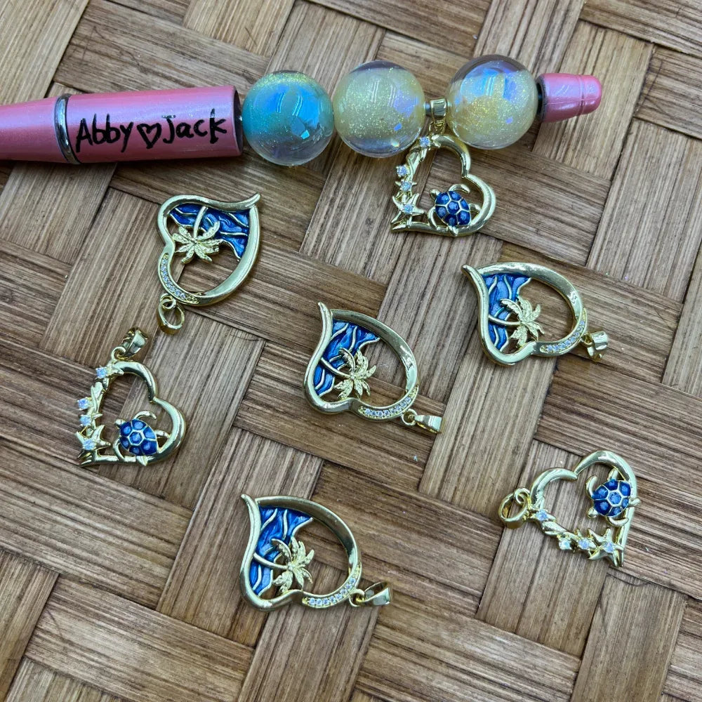 Gold tone enamel bail beads featuring blue ocean waves with palm tree design and sea turtle heart pendants, displayed on wicker surface next to iridescent globe beads and a beadable pen