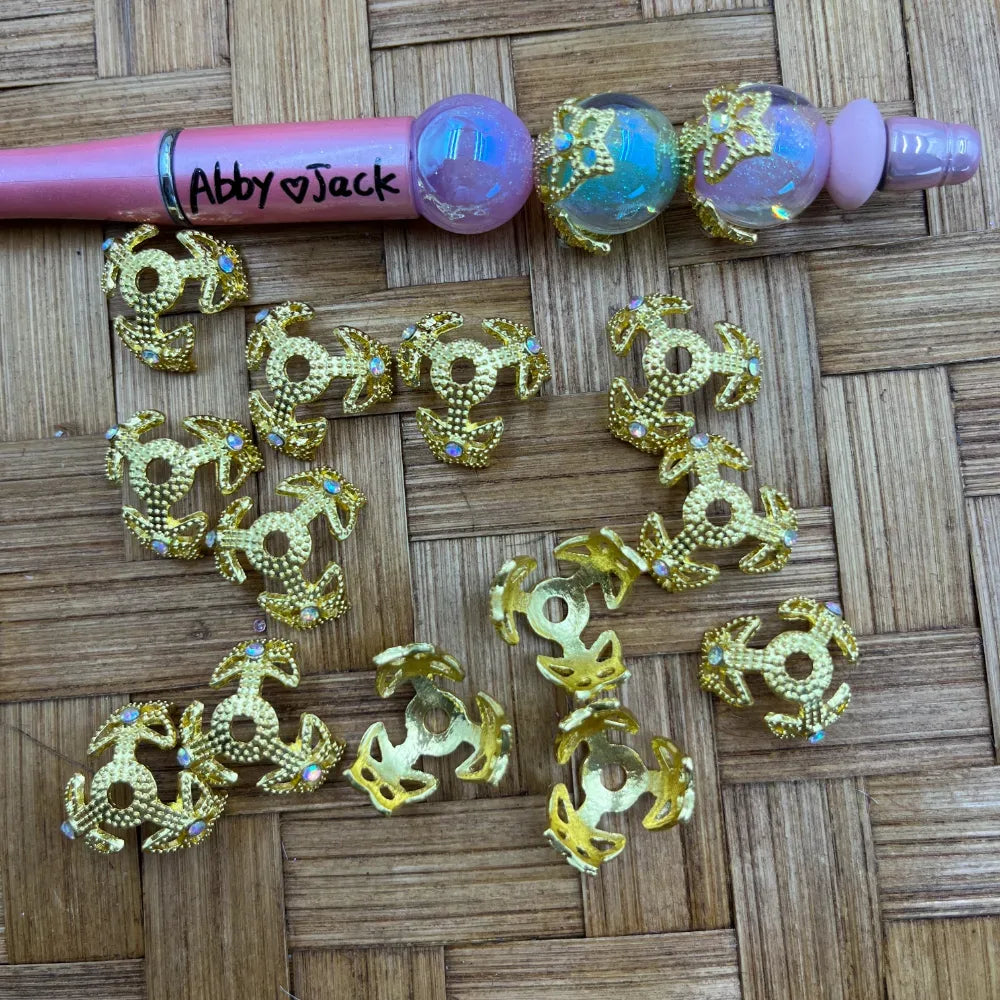 Gold filigree flower-shaped bead caps with iridescent AB rhinestone accents scattered on a woven surface next to a pink beadable pen labeled Abby Jack.