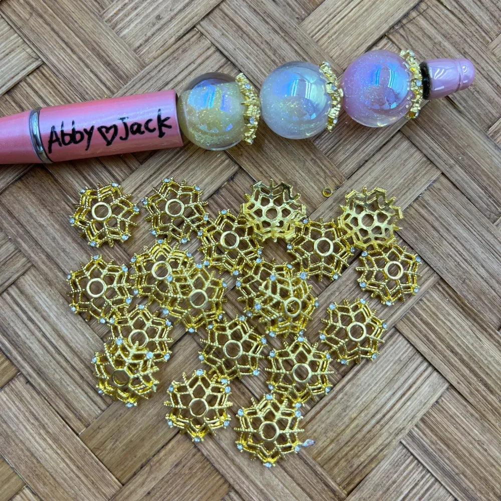 Gold filigree openwork flower bead caps with rhinestone accents displayed on woven surface next to beadable pens with globe beads, showing sparkly crystal detail points on each petal segment.