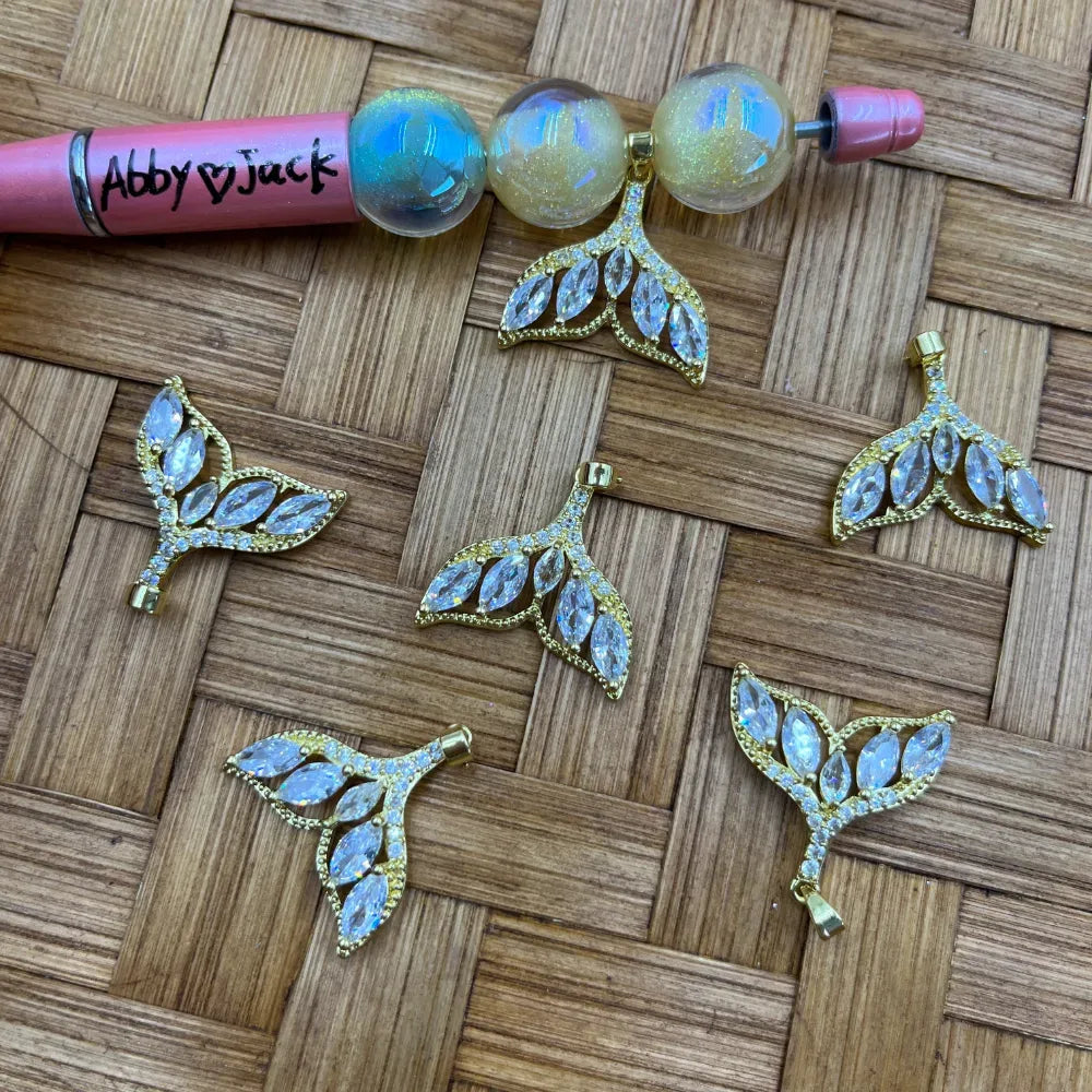 Five gold mermaid tail shaped bail beads with clear marquise CZ crystal rhinestones arranged on a wicker surface next to iridescent globe beads and a beadable pen.