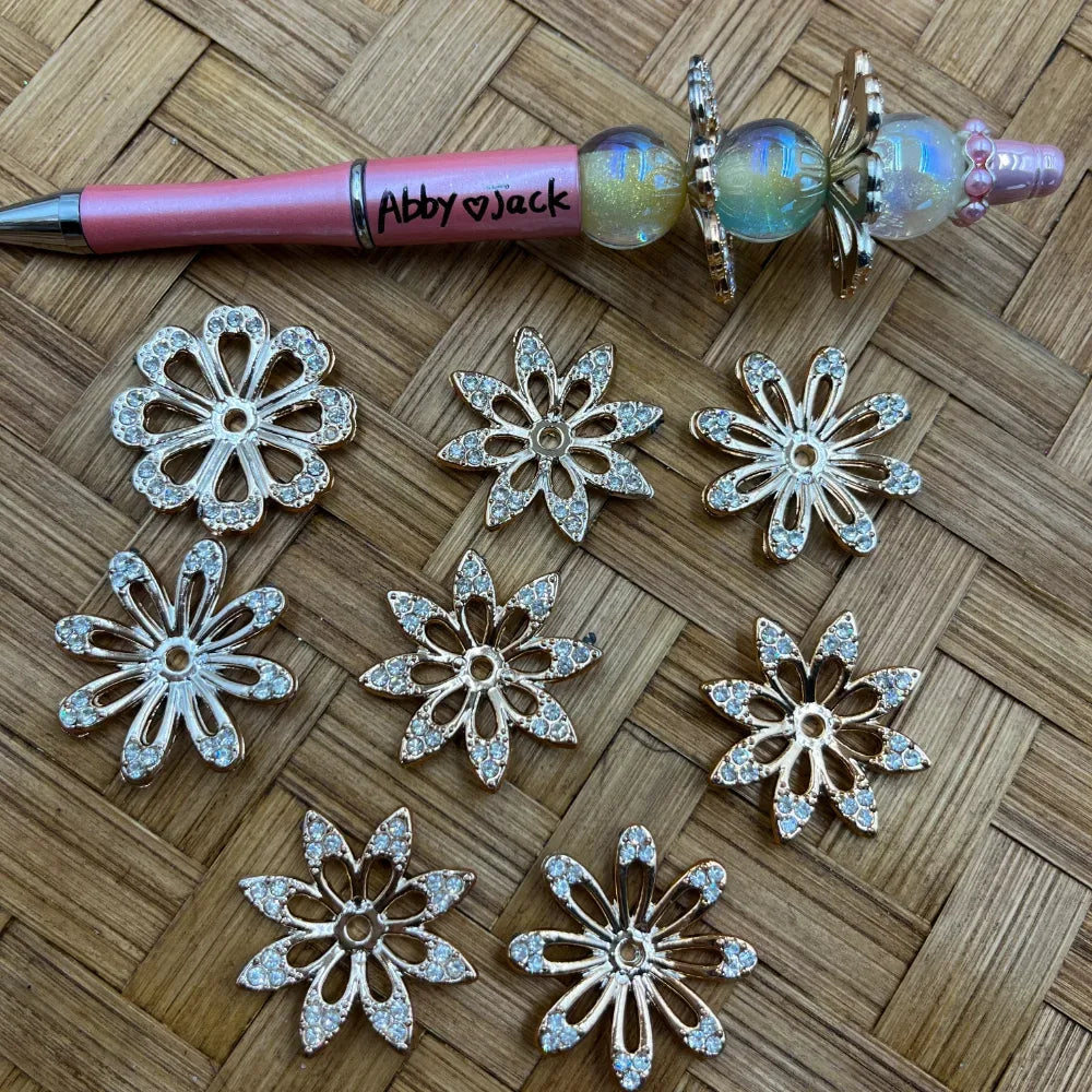 Gold metal rhinestone daisy flower spacer beads with crystal embellishments displayed next to a pink beadable pen on a wicker surface, sold at DIY Mini Planet.