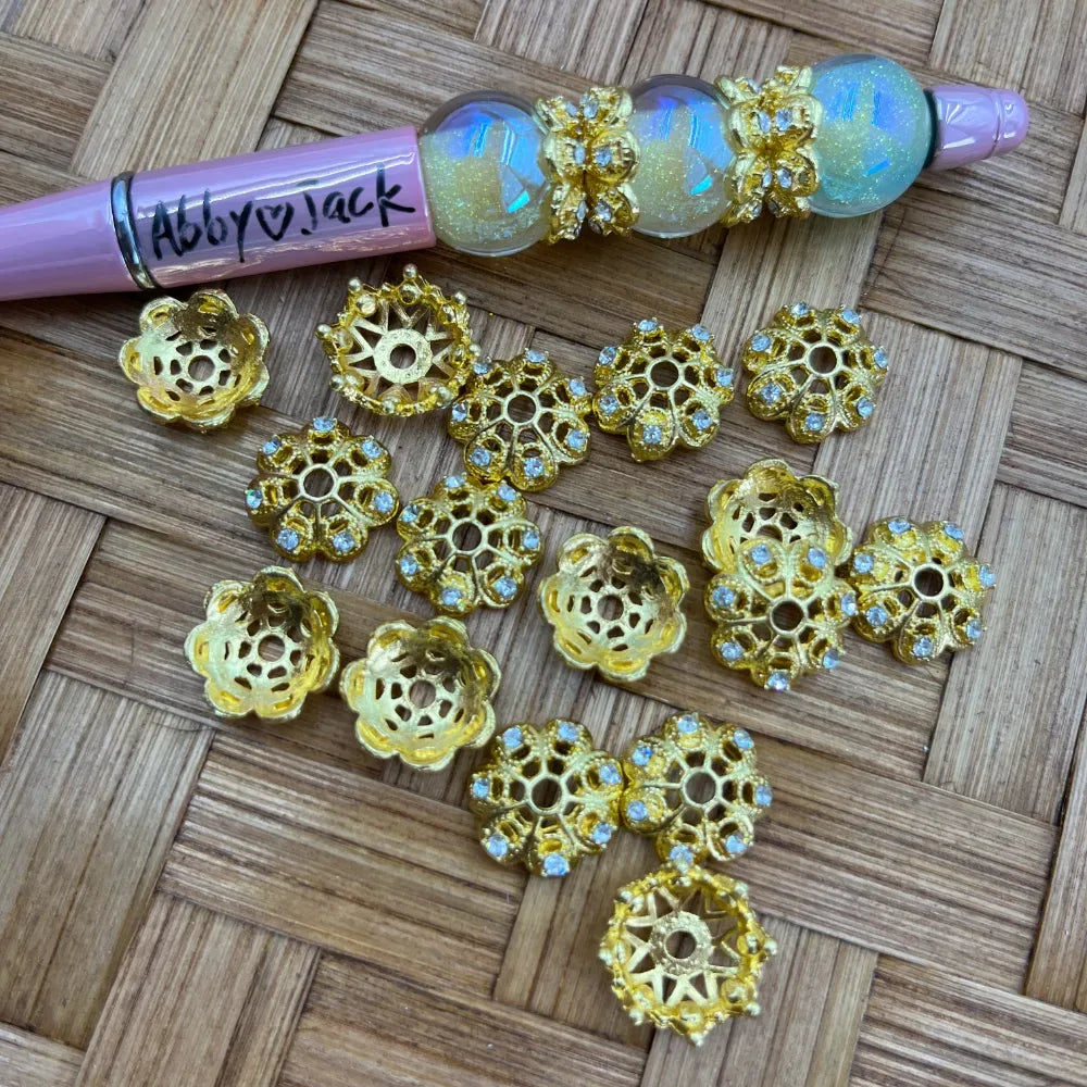 Assorted gold filigree flower bead caps with rhinestone crystal accents displayed on woven surface alongside a pink beadable pen decorated with iridescent beads and gold bead caps.