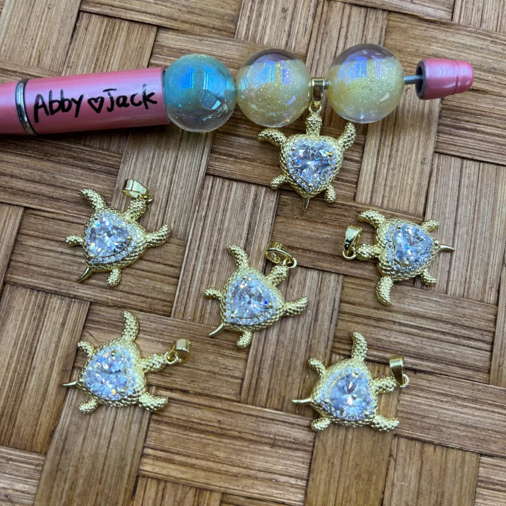 Six gold sea turtle charm pendants with heart-shaped CZ crystal shell centers arranged on a woven surface next to iridescent aurora borealis beads and a pink beadable pen labeled Abby and Jack.