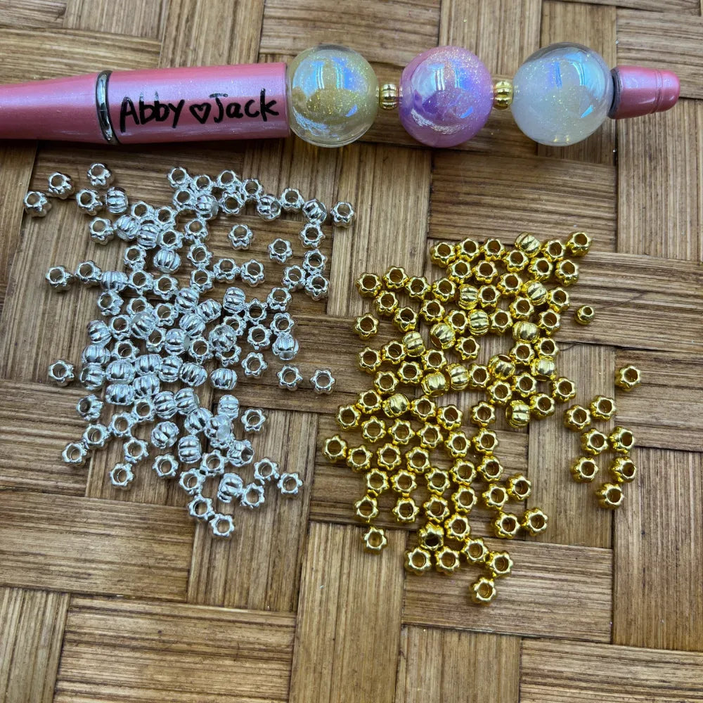 Pile of gold and silver corrugated ribbed metal spacer beads displayed on a woven surface next to a beadable pen decorated with iridescent globe beads, showing both color options for DIY jewelry making.