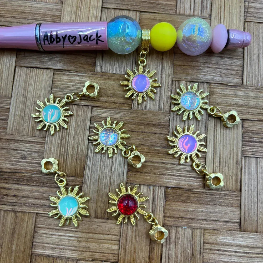 Gold sun-shaped charm bail beads with colorful iridescent and holographic cabochon centers in pink, teal, red, purple, and opal, displayed on a woven surface next to a decorated beadable pen.