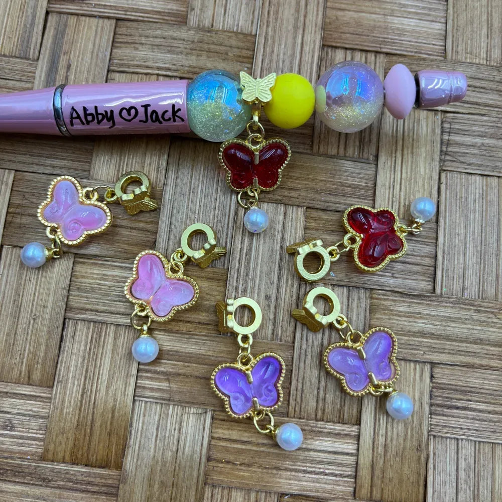 Assorted gold tone butterfly charm bail beads in pink, purple, and red with pearl dangles, displayed on a woven surface next to a decorated beadable pen labeled Abby and Jack.